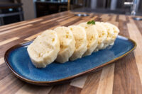 Czech Bread Dumpling Houskovy Knedlik Recipe Sliced houskový knedlík rounds on a blue plate, spongy crumb visible, garnished with parsley.