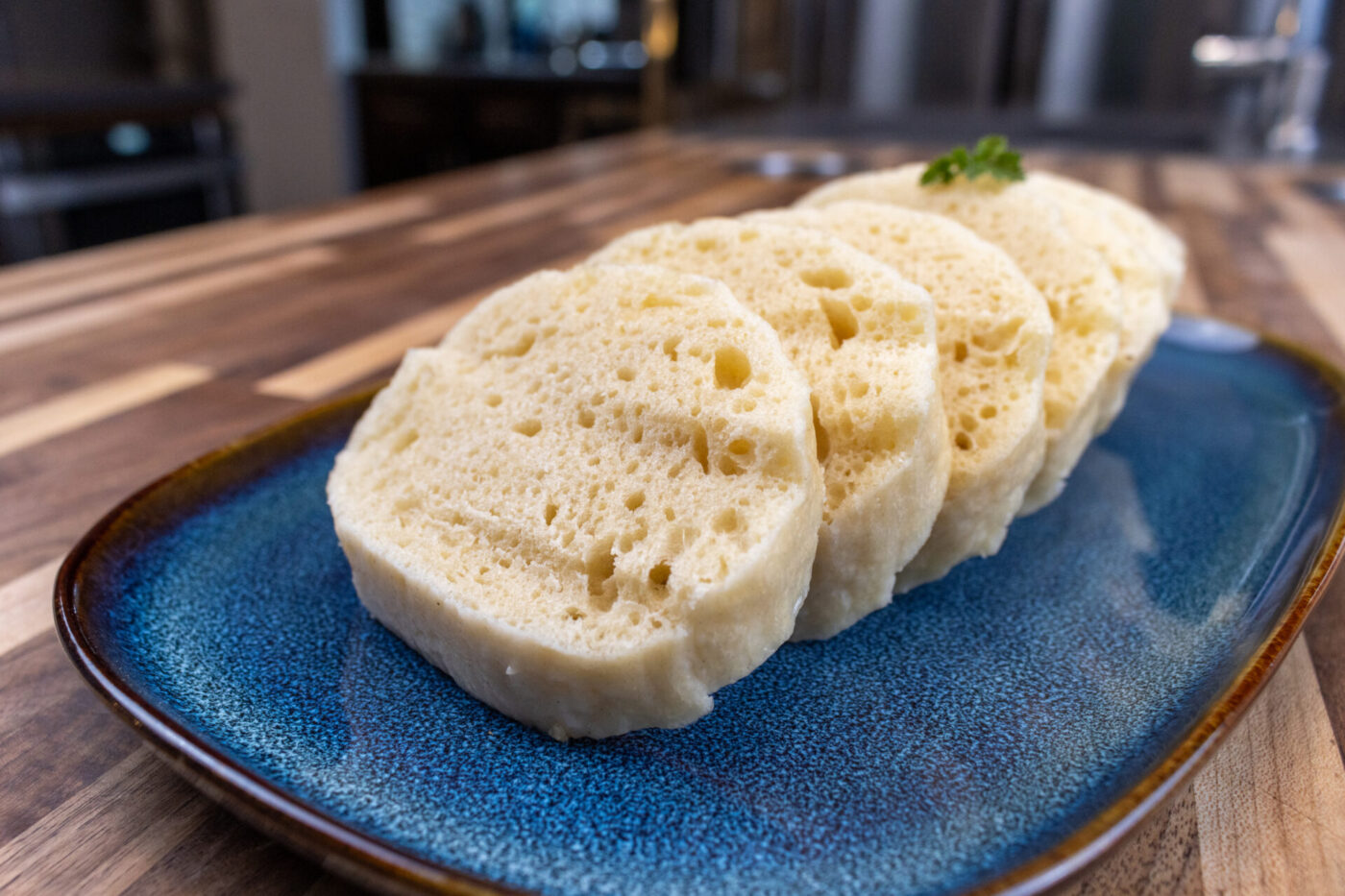 Czech Bread Dumpling Houskovy Knedlik Recipe Sliced houskový knedlík with visible air holes, garnished with herb, on a blue ceramic plate.