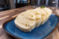 Czech Bread Dumpling Houskovy Knedlik Recipe Sliced houskový knedlík with visible air holes, garnished with herb, on a blue ceramic plate.