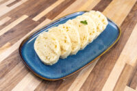 Czech Bread Dumpling Houskovy Knedlik Recipe Sliced houskový knedlík rounds with airy holes on a blue oval plate, garnished with a herb leaf.