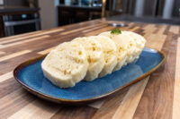 Czech Bread Dumpling Houskovy Knedlik Recipe Sliced houskový knedlík rounds fanned on a blue plate, showing spongy, porous texture, garnished with herb.