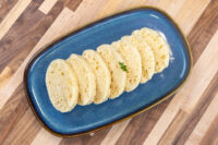 Czech Bread Dumpling Houskovy Knedlik Recipe Sliced houskový knedlík fanned on a dark blue plate, garnished with a small herb sprig.