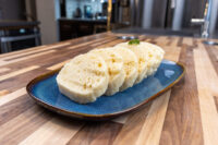 Czech Bread Dumpling Houskovy Knedlik Recipe Sliced houskový knedlík with a spongy, holed texture on a blue oval plate with herb garnish.