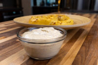 Glass bowl of Mustard Dipping Sauce for Soft Pretzels beside a plate of pretzel-topped bagels on a wood table.