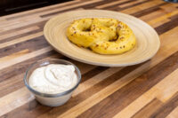 Glass bowl of Mustard Dipping Sauce for Soft Pretzels beside a seeded soft pretzel on a wooden plate.