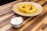 Glass bowl of Mustard Dipping Sauce for Soft Pretzels beside a pretzel bun on a wood table.