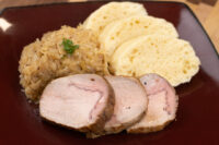 Houskový Knedlík With Pork Sliced houskový knedlík fanned beside sauerkraut and pork loin on a dark red plate.