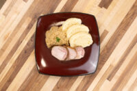 Houskový Knedlík With Pork Sliced houskový knedlík rounds beside pork loin and sauerkraut on a dark square plate