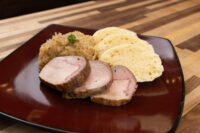 Sliced pork, sauerkraut, and dumplings arranged on a dark red plate for Vepřo-knedlo-zelo.