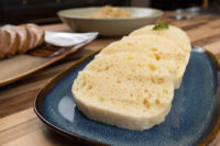 Czech Bread Dumpling Houskovy Knedlik Recipe Sliced houskový knedlík on a blue plate, with bread and a bowl blurred in the background