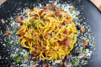Spaghetti Carbonara Twirled Spaghetti Carbonara on a black plate topped with crispy bacon bits, herbs and grated cheese.