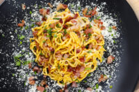Spaghetti Carbonara Overhead shot of Spaghetti Carbonara topped with crispy bacon, herbs, and grated cheese on a black plate.