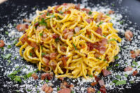 Spaghetti Carbonara Golden Spaghetti Carbonara twirled on a black plate with crispy bacon bits, herbs, and grated cheese.