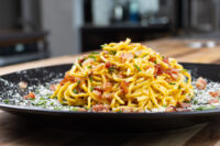 Spaghetti Carbonara Twirled Spaghetti Carbonara topped with crispy bacon, herbs, and grated cheese on a black plate.