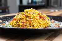 Spaghetti Carbonara Heaping plate of Spaghetti Carbonara with crispy bacon, herbs, and grated cheese on black plate.