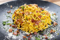 Spaghetti Carbonara Golden-sauced Spaghetti Carbonara topped with bacon bits, herbs, and grated cheese on a black plate.