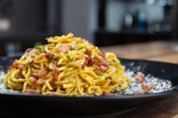 Spaghetti Carbonara Heaping plate of Spaghetti Carbonara with crispy bacon, chives, and grated cheese on black dish.
