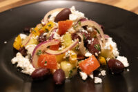 Greek Salad Greek Salad with diced peppers, tomato, red onion, kalamata olives and crumbled feta on a black plate.