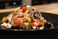 Greek Salad Black plate of Greek Salad with diced tomatoes, red onion, kalamata olives, and feta on a wooden table.