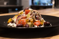 Greek Salad Greek Salad on a black plate with roasted vegetables, red onion, olives, and crumbled feta.