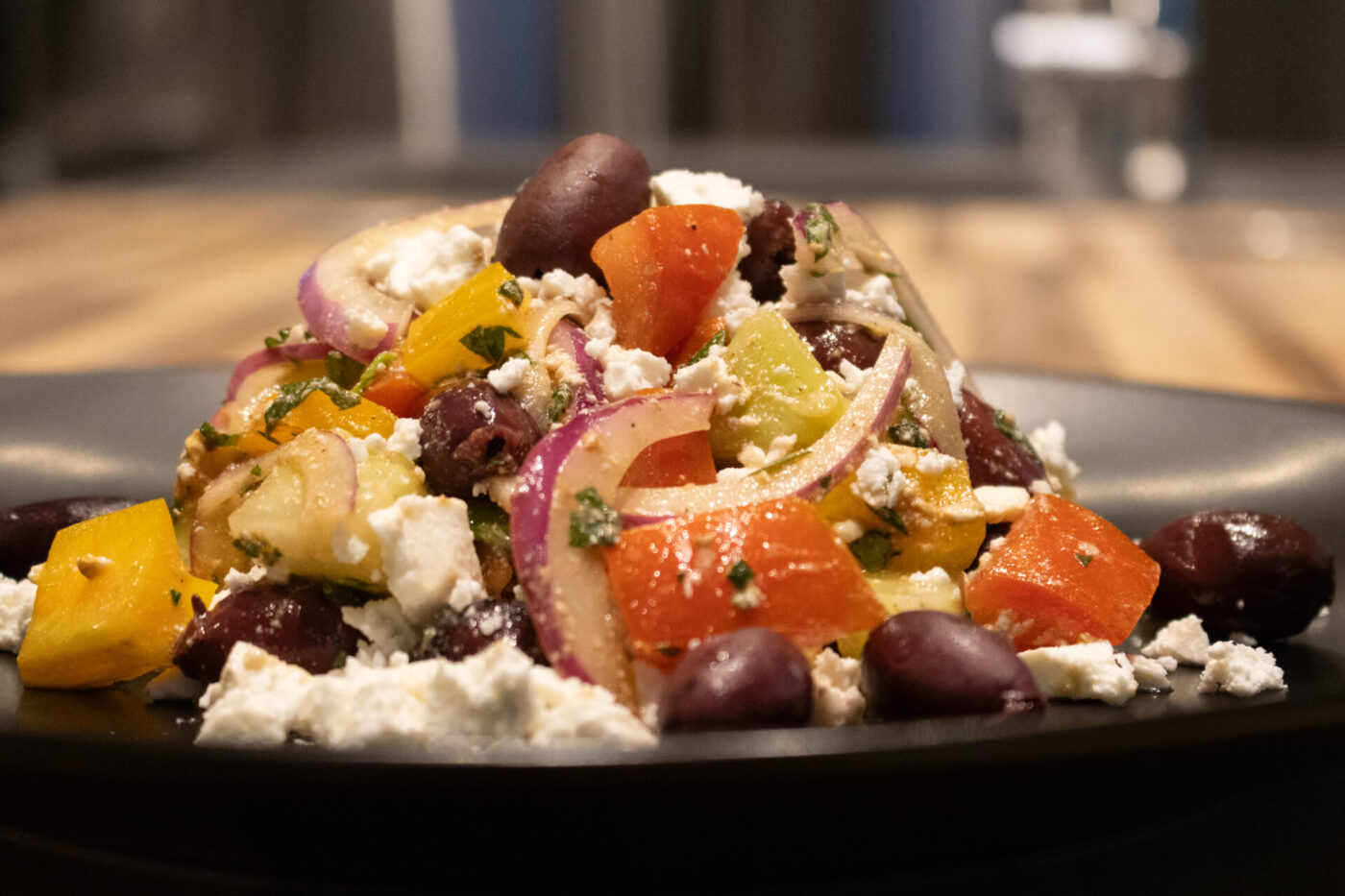 Greek Salad A mound of Greek Salad with diced peppers, red onion, kalamata olives, and crumbled feta on a dark plate.