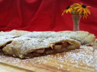 Apple Strudel Sliced Apple Strudel on a wooden board, dusted with powdered sugar, fruit filling visible in cross-section.