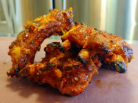 Czech BBQ – Honey Pork Ribs Glazed, charred Czech BBQ – Honey Pork Ribs pieces with a red-orange glaze on a light pink surface.
