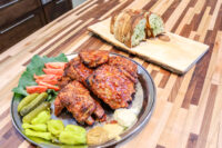 Czech BBQ – Honey Pork Ribs Glazed Czech BBQ – Honey Pork Ribs on a metal plate with pickles, tomatoes, greens, and mustard.