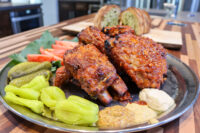 Czech BBQ – Honey Pork Ribs Glazed Czech BBQ – Honey Pork Ribs on a metal plate with pickles, peppers, tomato, and two dips.