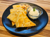 Triangular Quesadilla wedges on a black plate beside a glass bowl of sour cream with herb garnish