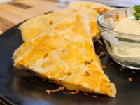 Golden quesadilla wedges with melted cheese filling and chili flakes on a black plate beside a bowl of sour cream.