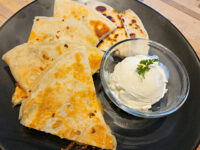 Crispy quesadilla wedges with charred spots on a black plate, next to a bowl of sour cream topped with parsley.