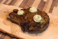 Cowboy Steak Bone-in Cowboy Steak with a charred crust, topped with three melting dollops of herb butter on a wooden board.