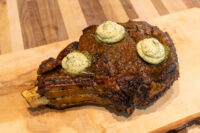 Cowboy Steak with Herb Butter Seared bone-in Cowboy Steak topped with three dollops of herb butter on a wooden board.