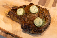 Cowboy Steak Bone-in Cowboy Steak with charred crust and three herb butter rounds melting on top, on a wooden board