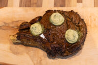 Cowboy Steak Bone-in Cowboy Steak with a dark seared crust, topped with three melting rounds of herb butter, on a wooden board.