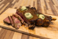 Cowboy Steak Bone-in Cowboy Steak topped with herb butter, partly sliced to show a pink center, on a wooden board