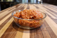 Italian Sausage Recipe Glass bowl of raw seasoned Italian Sausage mixture on a striped wooden countertop.