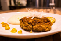 Karbenátky (Czech Burger) Breaded, fried Karbenátky (Czech Burger) patty on a white plate with mustard dots and a pickle slice.