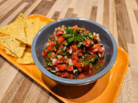 Grey bowl of chunky Salsa with diced tomatoes, onion, and herbs, tortilla chips on an orange tray.