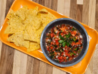 Bowl of chunky Salsa with diced tomato and herbs next to tortilla chips on an orange tray