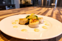 Scallops with Spicy Sauce Three seared scallops with lemon and greens on a white plate, dotted with pale sauce, for Scallops with Spicy Sauce.