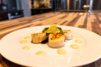 Scallops with Spicy Sauce Four seared scallops with a lemon slice, arugula, and dots of spicy sauce on a white plate.