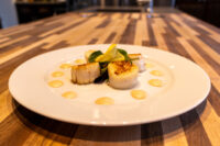 Scallops with Spicy Sauce Seared scallops with spicy sauce dots, arugula, and lemon garnish on a white plate on wood table.