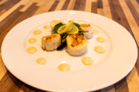 Scallops with Spicy Sauce Four seared scallops with lemon, arugula, and dots of spicy sauce on a white plate