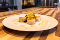 Scallops with Spicy Sauce Three seared scallops on a white plate with lemon wedge, greens, and dots of pale sauce