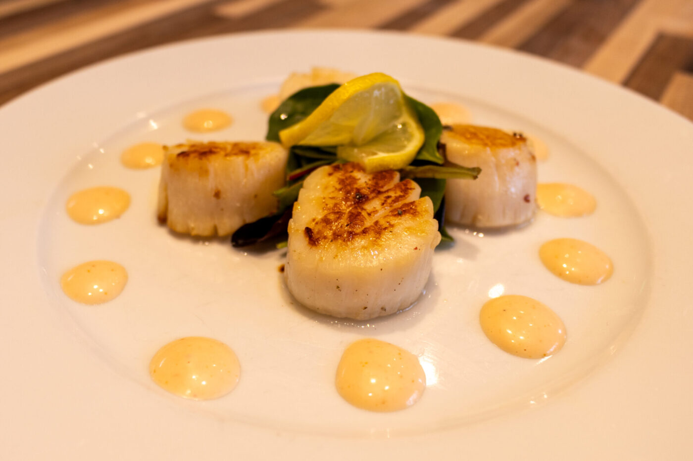 Scallops with Spicy Sauce Three seared scallops with lemon twist and arugula, dots of spicy sauce ringing the plate