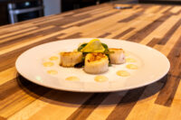 Scallops with Spicy Sauce Three seared scallops with pale sauce dots, lemon and arugula garnish, Scallops with Spicy Sauce on a white plate.