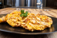 Almond Chicken Schnitzel Golden Almond Chicken Schnitzel topped with sliced almonds, garnished with parsley on a black plate.