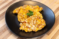 Almond Chicken Schnitzel Almond Chicken Schnitzel coated in sliced almonds, garnished with parsley, on a black plate.